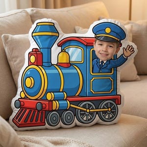 Peut inclure: Un coussin fantaisie en forme de train de dessin animé, avec un enfant souriant en uniforme de conducteur saluant de la fenêtre. Le train est principalement bleu avec des détails rouges et jaunes. Le coussin est sur un canapé beige.