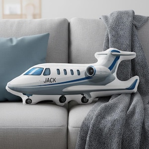 Peut inclure: Un coussin en forme d'avion blanc et bleu avec le nom "JACK" imprim&eacute; sur le c&ocirc;t&eacute;. Le coussin a des accents bleus et repose sur un canap&eacute; gris avec un coussin bleu et une couverture grise.