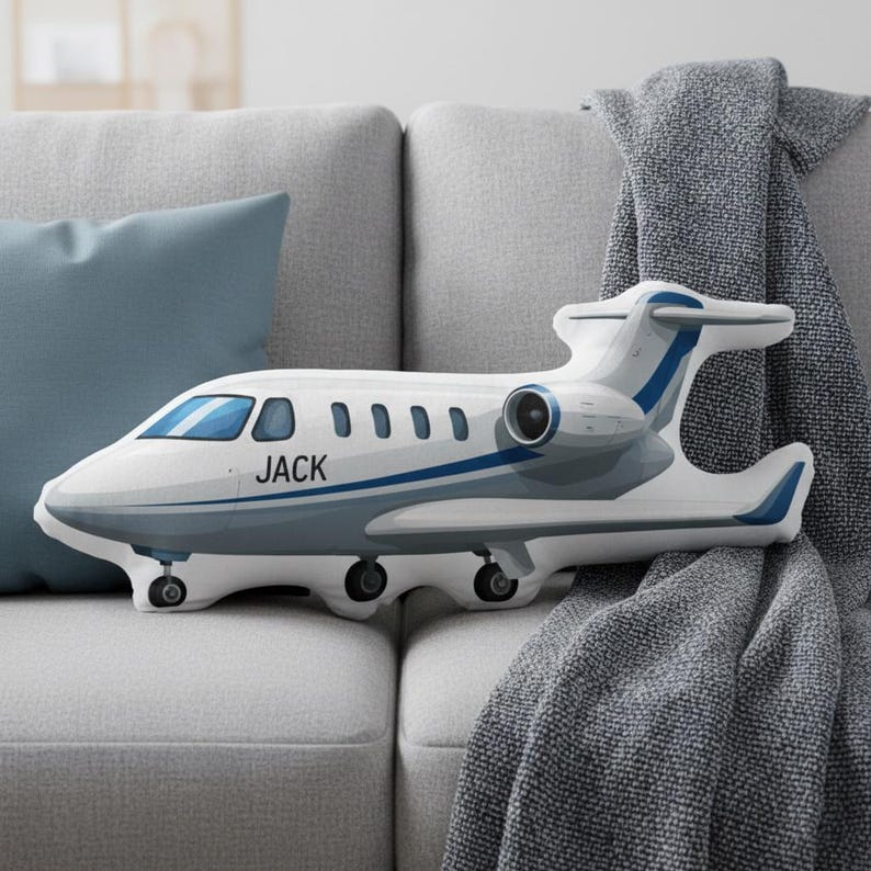 Peut inclure: Un coussin en forme d'avion blanc et bleu avec le nom "JACK" imprim&eacute; sur le c&ocirc;t&eacute;. Le coussin a des accents bleus et repose sur un canap&eacute; gris avec un coussin bleu et une couverture grise.