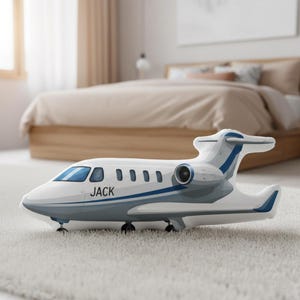 Peut inclure: Un avion jouet blanc et bleu avec le nom "JACK" imprim&eacute; sur le c&ocirc;t&eacute;. Le jet jouet pr&eacute;sente un design d&eacute;taill&eacute;, comprenant des fen&ecirc;tres, un r&eacute;acteur et un empennage. Il repose sur un tapis blanc.