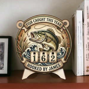 Puede incluir: Letrero de madera con temática de pesca con una ilustración de lubina. El letrero dice "FISH CAUGHT THIS YEAR" y "HOOKED BY JAMES", con un contador que muestra "162". El letrero se muestra en un soporte de madera.