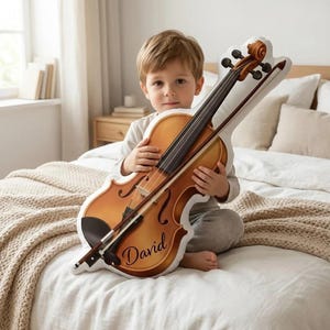 May include: A plush violin-shaped pillow in shades of brown and black, with a bow. The pillow has the name "David" printed on it. The child is holding the pillow on a bed with a white comforter and a beige blanket.