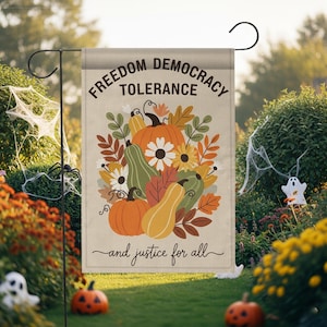 May include: A beige garden flag with the words "Freedom Democracy Tolerance" and "and justice for all." The flag features a colorful illustration of pumpkins, gourds, leaves, and flowers. The flag is hanging in a garden setting with Halloween decorations.
