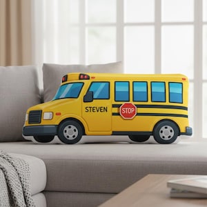 May include: A yellow school bus-shaped pillow with black wheels and trim. The pillow has blue windows and the name "STEVEN" printed on the side. A red "STOP" sign is also visible.