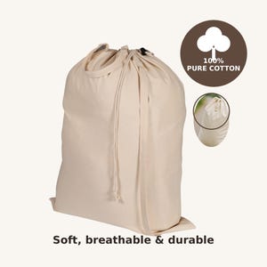 Puede incluir: Una bolsa de lavandería de algodón de color natural con cierre de cordón. La bolsa está etiquetada como "100% Pure Cotton" y "Soft, breathable & durable". La bolsa está diseñada para guardar la ropa sucia y tiene un diseño sencillo y funcional.