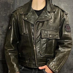 May include: Dark green leather jacket with a classic biker style. Features a notched collar, multiple pockets, and a zip-up front. Includes patches on the sleeves and chest, and a belt at the waist. The jacket has a distressed, vintage look.