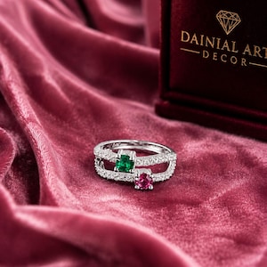 Peut inclure: Une bague en argent avec deux rangées, l'une au-dessus de l'autre, incrustées de petites pierres claires. La bague présente un émeraude carrée et un rubis carré. La bague est présentée sur une surface de velours rose.