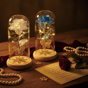 Op de afbeelding: Twee decoratieve glazen koepels met LED-rozen, één iriserend, de andere blauw. Elke koepel staat op een houten voet met een vlinder en bloemenmotief. Op een houten oppervlak liggen een rode roos en een parelketting.