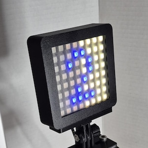 Puede incluir: Una pantalla LED matricial negra con una cuadrícula de luces blancas, amarillas y azules. Las luces forman la letra "R". La pantalla está montada sobre un soporte negro con tornillos.