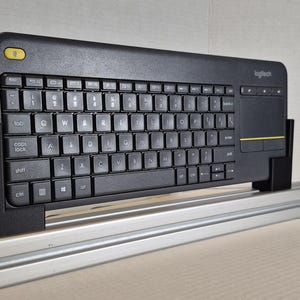 Może przedstawiać: Czarna bezprzewodowa klawiatura Logitech z wbudowanym panelem dotykowym. Klawiatura ma pełny układ QWERTY z białymi literami na klawiszach. Żółty przycisk znajduje się w lewym górnym rogu. Klawiatura jest zamontowana na metalowej szynie.