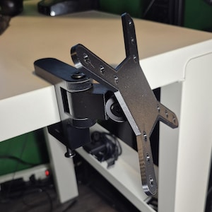 Könnte beinhalten: Ein schwarzer Monitorhalter, der an einem weißen Schreibtisch befestigt ist. Die Halterung hat einen schwenkbaren Arm und eine VESA-Platte mit mehreren Befestigungslöchern. Der Schreibtisch ist weiß, der Hintergrund unscharf.