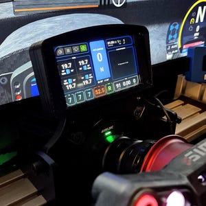 May include: A black Simagic racing steering wheel display unit featuring a digital screen with telemetry data, including tire pressures of 19.7, a pit limiter set to 0, and a neutral gear indicator. The device has a green LED light and red accents.