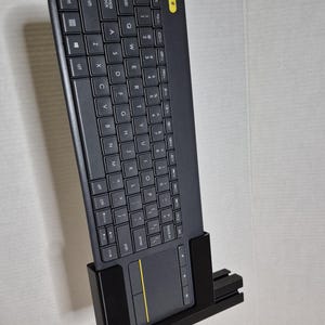 Op de afbeelding: Een zwart draadloos toetsenbord met een ingebouwd touchpad en een gele accentknop. Het toetsenbord is gemonteerd op een zwarte standaard. Het toetsenbord heeft een standaard QWERTY-indeling.