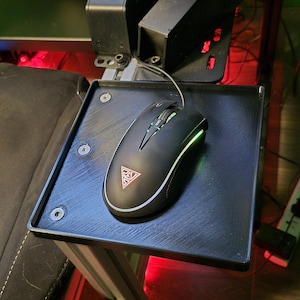 May include: A black gaming mouse with a white trim and a triangular logo, resting on a black, square platform. The platform is attached to a metal frame, and the mouse has a green illuminated scroll wheel and side lights. The setup suggests a gaming or simulation environment.