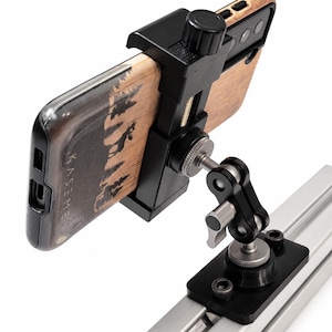 May include: A black smartphone holder with a silver and black adjustable arm, mounted on a silver rail. The phone is in a wooden case with a forest and moose design. The holder has a black knob for adjustment.