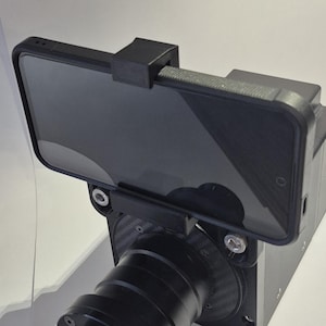 May include: A black smartphone is mounted on a black camera rig. The phone is held in place by a black bracket. The camera rig has a black lens and a black body. The setup is designed for video recording.