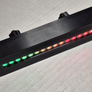 May include: A black rectangular device with a row of multi-colored LED lights. The lights transition from green to orange to red. The device has two mounting brackets and a cable.