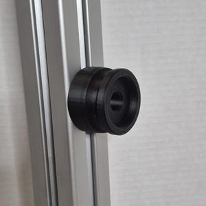 May include: A black, circular 3D-printed component attached to a silver metal extrusion. The component has a central hole and a ridged outer edge. The metal extrusion has vertical grooves.