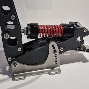 Puede incluir: Primer plano de un conjunto de pedales de carreras negro y plateado con un resorte rojo. El conjunto presenta una base plateada, un marco negro y varios pernos y tornillos. El resorte rojo está posicionado dentro del marco negro.