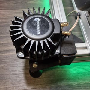 Op de afbeelding: Een zwart Dayton Audio luidsprekercomponent met een stekelig, ventilatorachtig ontwerp. Het component is gemonteerd op een zwarte beugel met goudkleurige connectoren en een draad. De luidspreker staat op een houten oppervlak met groene verlichting.