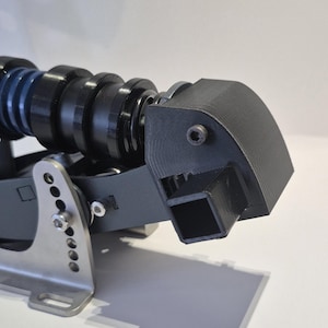 Könnte beinhalten: Nahaufnahme eines schwarzen und silbernen mechanischen Geräts. Das Gerät verfügt über eine Reihe von schwarzen zylindrischen Komponenten, ein gebogenes schwarzes Gehäuse und eine silberne Metallbasis mit mehreren Löchern und einer Schraube. Das Gerät befindet sich vor einem weißen Hintergrund.