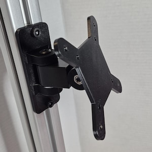 Puede incluir: Soporte de monitor negro y ajustable, fijado a un marco de metal plateado. El soporte tiene un brazo giratorio y una placa cuadrada con múltiples orificios para tornillos. Diseño funcional e industrial.