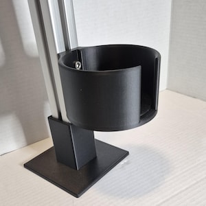 Puede incluir: Un portavasos negro fijado a un soporte de metal plateado y negro. El portavasos es cilíndrico y tiene una base plana. El soporte tiene una base cuadrada y un soporte vertical. El soporte mide aproximadamente 25 cm de altura.