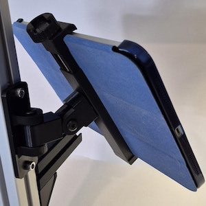 May include: A black adjustable tablet mount attached to a vertical silver metal rail. The mount features a spring-loaded clamp holding a tablet with a blue cover, secured by a black knob at the top and a multi-jointed arm for positioning.