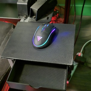 May include: A black gaming mouse with a glowing RGB light sits on a black pull-out tray. The mouse has a triangular logo and a curved design. The tray is part of a larger black structure, possibly a gaming setup.