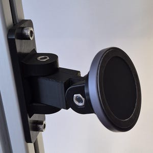 May include: A black, adjustable phone mount attached to a silver metal frame. The mount features a circular, magnetic head and is secured with bolts. The design is modern and functional, suitable for various devices.