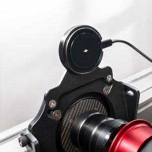 Puede incluir: Primer plano de un cargador inalámbrico negro con un símbolo de rayo, montado en un soporte de metal negro. El soporte está fijado a un dispositivo con un componente de fibra de carbono y rojo. Un cable negro se extiende desde el cargador.