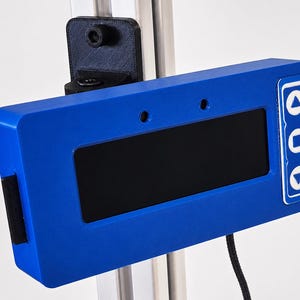 May include: A blue rectangular device with a black screen and white control buttons. The device is mounted on a metal frame with a black bracket. A black cable extends from the bottom. The device has a small black strip on the side.