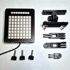 May include: A black electronic device with a grid of white squares, a carbon fibre texture, and a connecting cable. Additional black mounting hardware and screws are also present, suggesting a tech accessory or component.