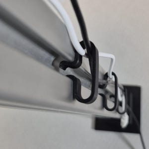 May include: Close-up of a metal track with black and white hooks holding black and white wires. The track is silver and the hooks are made of plastic. The wires are thin and appear to be electrical cords.
