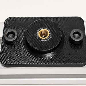 May include: A black, rectangular mounting plate with a central threaded insert and two hex head bolts. The plate is textured and sits on a silver metal rail. The threaded insert is gold in color.
