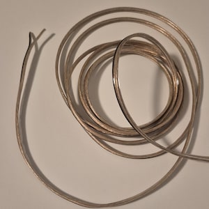 May include: Coiled silver-colored speaker wire with two exposed wire ends. The wire is looped in a circular pattern, suggesting it is new and ready for use. The image is taken on a white background.