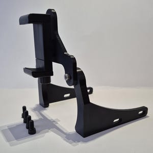 May include: A black metal device with a clamp, a pivoting arm, and a base with mounting holes. Several black bolts are placed near the base. The device appears to be a mounting bracket or holder for an electronic device.