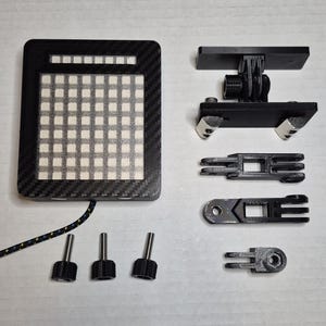 May include: A black and gray electronic device with a grid of white squares, a carbon fiber texture, and a braided cable. Additional black components and screws are also present, likely for mounting or adjustment.