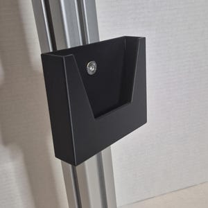 May include: A black, rectangular wall-mounted holder with a sloped front and a small, circular fastener. The holder is attached to a vertical, silver-coloured metal support. The background is a light, textured wall.