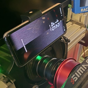 May include: A smartphone mounted on a black and red racing simulator component. The phone displays the time as 12:45. The simulator has a black base with red and green accents, and a blue display.