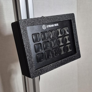 Può includere: Un Stream Deck nero con 15 pulsanti illuminati. Il dispositivo è montato su un telaio metallico. Il testo "STREAM DECK" è visibile nella parte superiore del dispositivo.