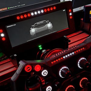 May include: A black SIMMAGIC racing wheel with red and white LED lights. The wheel has multiple buttons, dials, and a screen displaying a car graphic. The screen reads "ENDURANCE V1.0.0". A small screen displays "RACELOG".
