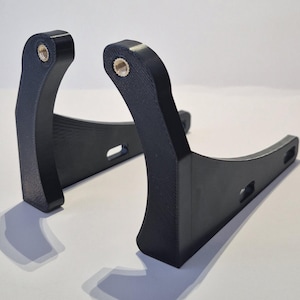 May include: Two black, curved brackets with threaded inserts at the top. The brackets have a sturdy, angular design with a curved base and a rectangular cutout. The items are likely for mounting or support.