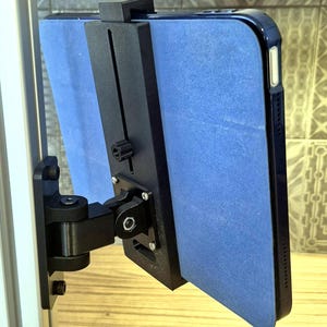 May include: A black tablet mount with a blue tablet case attached. The mount is made of black plastic and has several adjustable joints. The tablet is held securely in place by the mount, which is attached to a vertical surface.