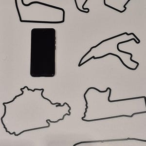 May include: Black outline designs of various race tracks, including the Nürburgring, with a black smartphone for size comparison. The tracks are displayed on a white surface.