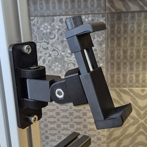 May include: A black adjustable phone mount attached to a metal frame. The mount features a pivoting arm and a clamp to secure a phone. The mount is designed to hold a phone in place.