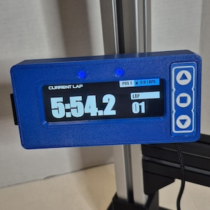 Puede incluir: Un dispositivo electrónico azul muestra "CURRENT LAP" y un tiempo de "5:54.2". El dispositivo también muestra "POS 1" y "0.0 LAPS". Tiene botones laterales y está montado en un marco de metal. El contador de vueltas indica "LAP 01".