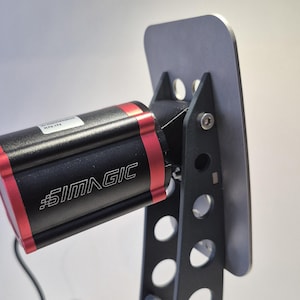 Puede incluir: Primer plano de un dispositivo SIMAGIC negro y rojo. El dispositivo tiene un cuerpo negro rectangular con detalles en rojo y el logotipo de SIMAGIC en blanco. Está montado en un marco de metal negro con una placa plateada.