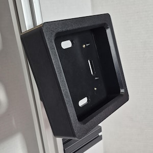 Puede incluir: Un soporte negro de forma cuadrada con superficie texturizada. Tiene dos orificios ovalados y una ranura vertical. El soporte está montado en un marco de metal plateado, mostrando su diseño y funcionalidad.