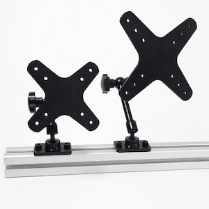 May include: Two black metal monitor mounting brackets with X-shaped plates attached to a silver aluminum T-slot rail. Each bracket features an adjustable arm with a black plastic tightening knob and a rectangular base plate secured to the rail.